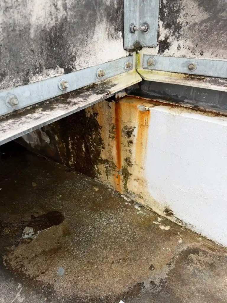 Water tank base corner with dark surface staining, rust marks and damp concrete below